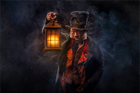 Giles Terera as Ebenezer Scrooge in A Christmas Carol at The Old Vic, holding a glowing lantern in swirling smoke. Photo by Hugo Glendinning