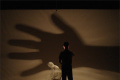 A figure stands beside a small sculpture against an enormous projected hand shadow in Andrew Dawson's Absence and Presence. Photo by Steve Smith