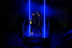 Three performers between two vertical blue strip lights on the set of WITNESS. Photo by Adrianne McKenzie