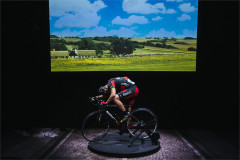 Connor Delves riding beneath a projection of open countryside in Cadel: Lungs On Legs at Riverside Studios. Photo by Kyle Dax Smith