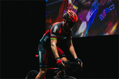 Connor Delves on the bike in front of a colourful projection in Cadel: Lungs On Legs at Riverside Studios. Photo by Kyle Dax Smith