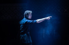 A man points into the darkness in a shaft of blue light in Martin Guerre. Photo by Helen Maybanks