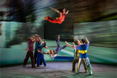 A performer in red is thrown high into the air above the company in front of projected landscapes in Motionhouse's Hidden