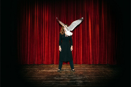 A performer stands before a red stage curtain, her face obscured by a gull's outstretched wings, in NINA at Battersea Arts Centre. Photo by Aimee Capstick