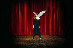 A performer stands before a red stage curtain, her face obscured by a gull's outstretched wings, in NINA at Battersea Arts Centre. Photo by Aimee Capstick