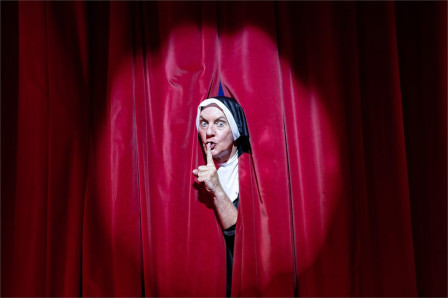 A performer in a nun's habit peers through red stage curtains with a finger to their lips, in The Pleasures of Fanny at Brick Lane Music Hall
