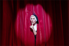 A performer in a nun's habit peers through red stage curtains with a finger to their lips, in The Pleasures of Fanny at Brick Lane Music Hall