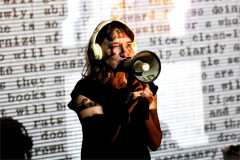 Emily Woof speaking into a megaphone against a wall of projected typescript in Revolver. Photo by Sheila Burnett
