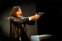 Emily Woof in a leather jacket, aiming a revolver in Revolver. Photo by Sheila Burnett