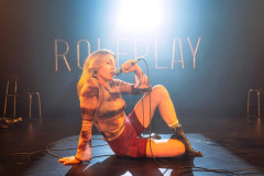 Hannah Reilly sits on the stage floor singing into a microphone beneath the neon ROLEPLAY sign in Roleplay at Soho Theatre