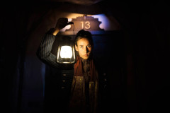 A performer in a long scarf holds up a lit lantern beneath a door marked 13 in Room 13 at Battersea Arts Centre