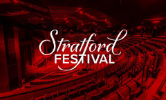 The auditorium of the Stratford Festival Theatre, its thrust stage and curved rows of empty seats lit in deep red, with the Stratford Festival logo across the centre