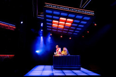 Two figures lean on a counter with drinks and snacks beneath a ribbon board reading LET’S GO CLIPPERS in Table 17 at Kiln Theatre. Photo by Steve Gregson