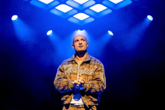 Jamael Westman as Dallas stands alone under blue light with his hands clasped in Table 17 at Kiln Theatre. Photo by Steve Gregson