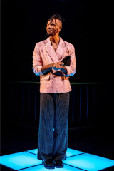 Michael Rishawn in a pink marbled blazer and pinstriped trousers, standing on the lit floor in Table 17 at Kiln Theatre. Photo by Steve Gregson