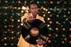 A performer in a cream suit holds out a vinyl record labelled The Brown Suite in front of a wall of records