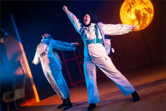 Two performers in white flight suits celebrate beneath a glowing planet in Toto at Polka Theatre