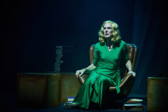 A performer in a green dress seated in an armchair in The Law of Mayhem