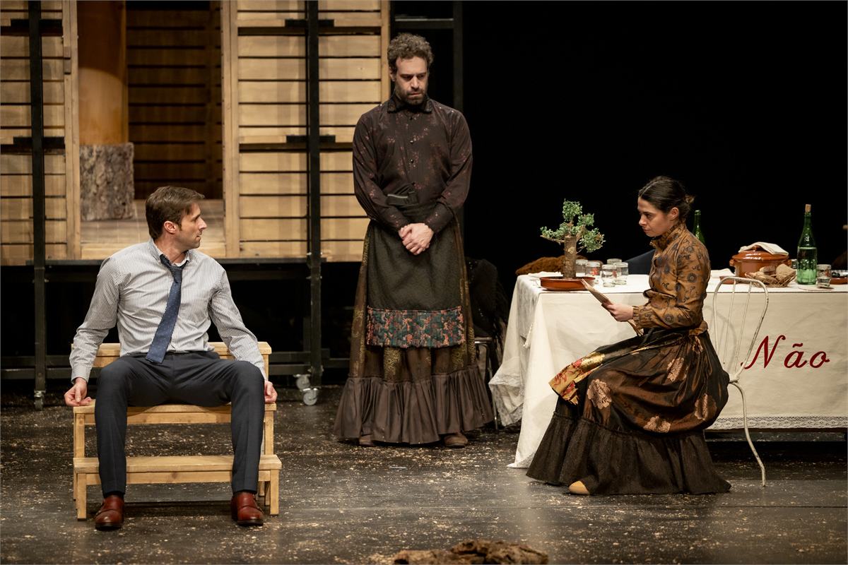 The kidnapped speechwriter sits on a wooden stool as two of the Catarinas watch him in Catarina and the Beauty of Killing Fascists