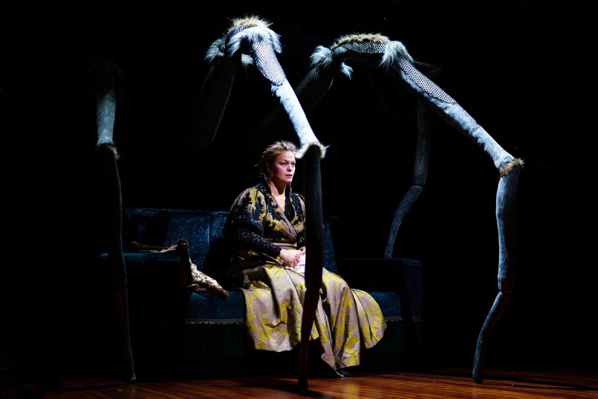 A performer in period dress sits on a sofa beneath towering puppet spider legs in Plexus Polaire's A Doll's House. Photo by Johan Karlsson