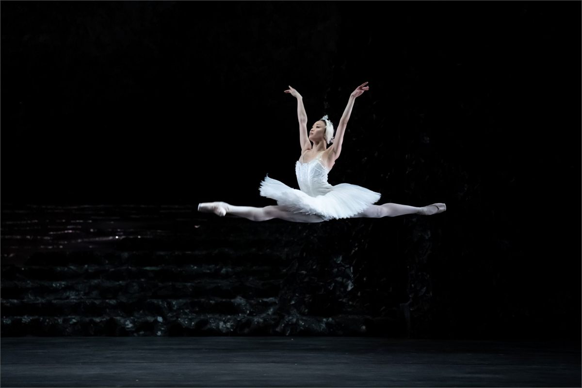 A ballerina in a white tutu leaps across a dark stage in Birmingham Royal Ballet's Swan Lake
