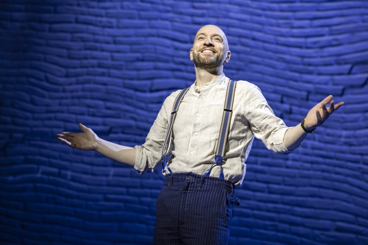 Derren Brown in braces and a white shirt, arms outstretched against a blue backdrop, in Only Human at the Apollo Theatre