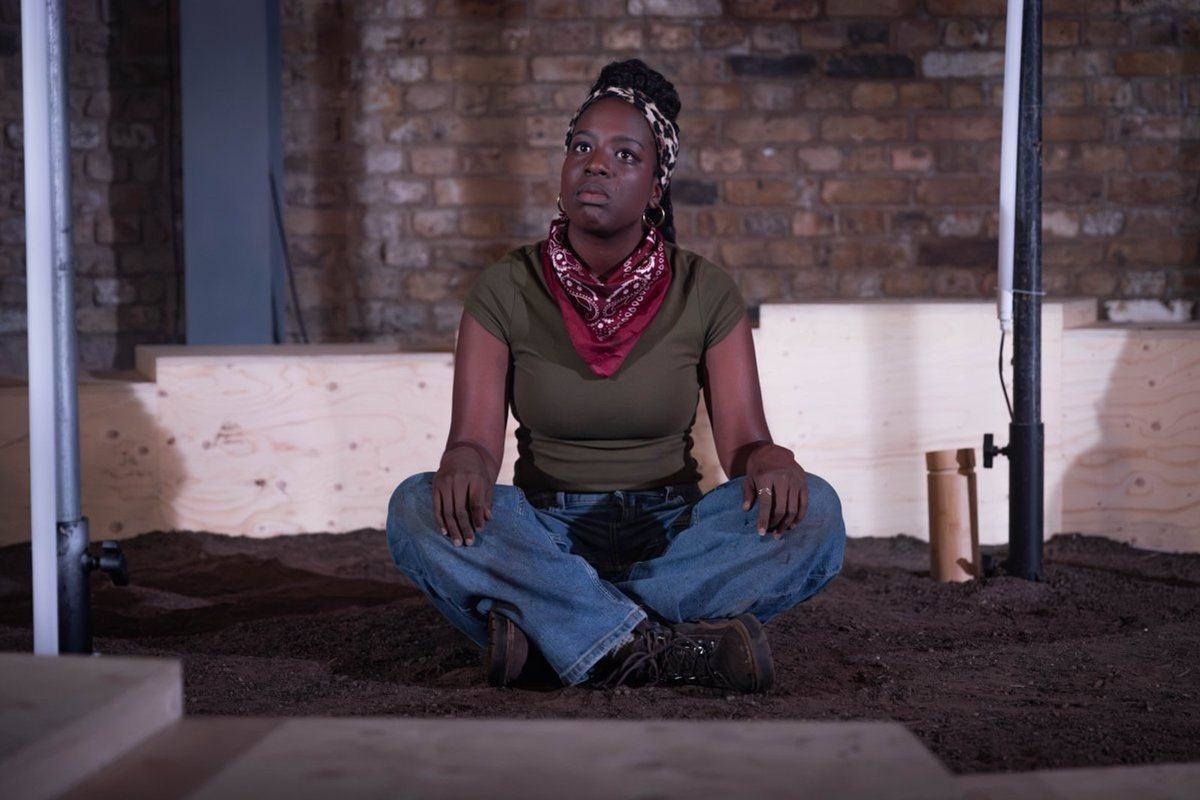 Gabrielle Brooks as Paige sits on the earth of the set in Fire Fire at the Arcola Theatre