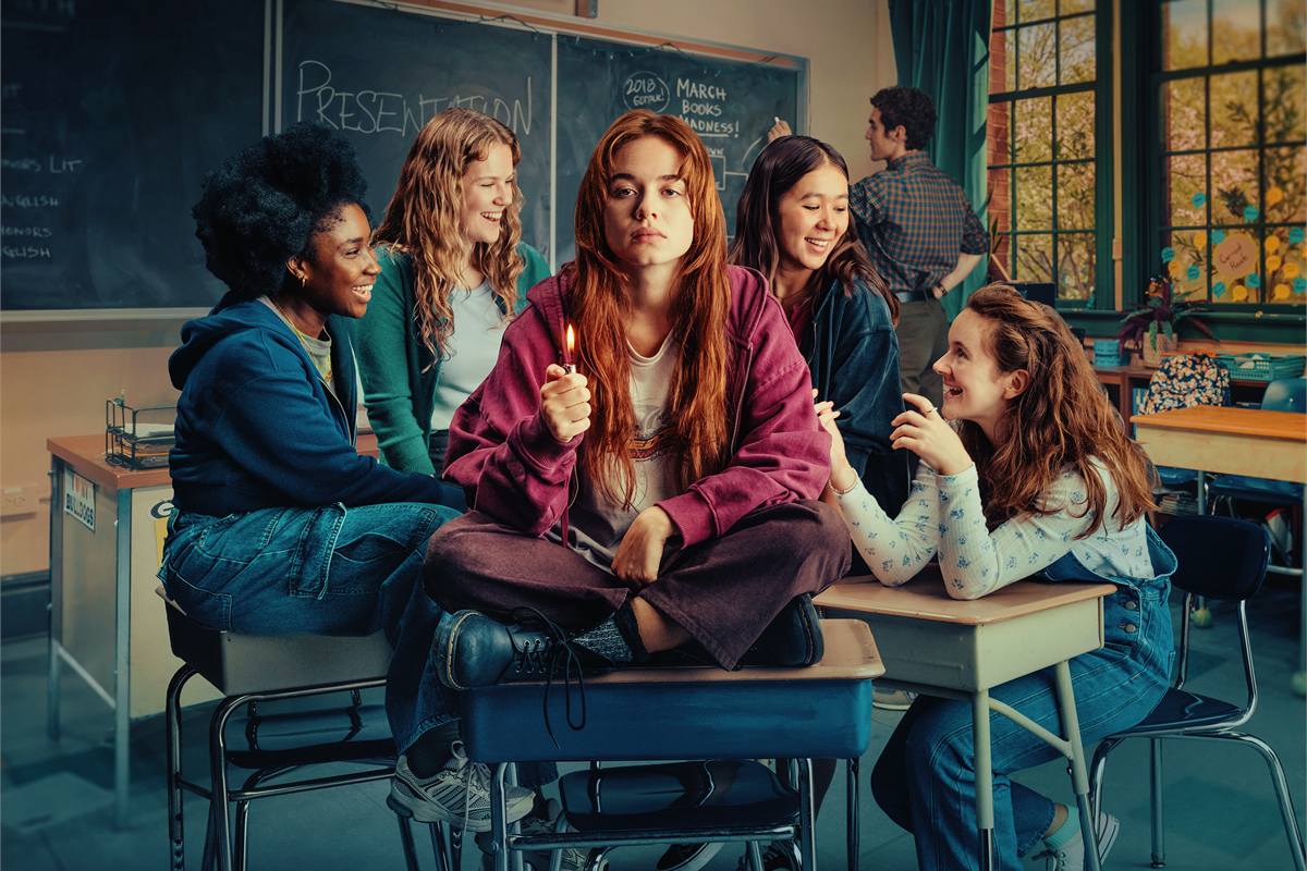 John Proctor Is the Villain artwork: a young woman holding a lit match sits cross-legged on a school desk, surrounded by classmates. Photo by Camilla Greenwell