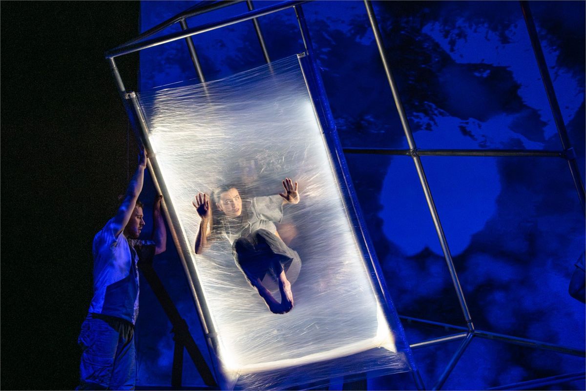 A performer is wrapped in a sheet of plastic inside a metal frame under blue light in Motionhouse's Hidden