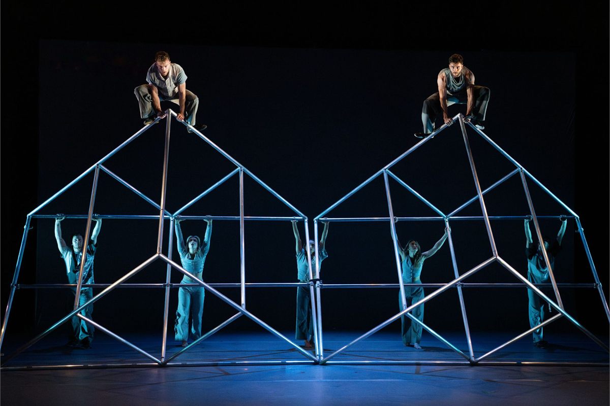 Two performers crouch on top of two metal frames while others stand inside them in Motionhouse's Hidden