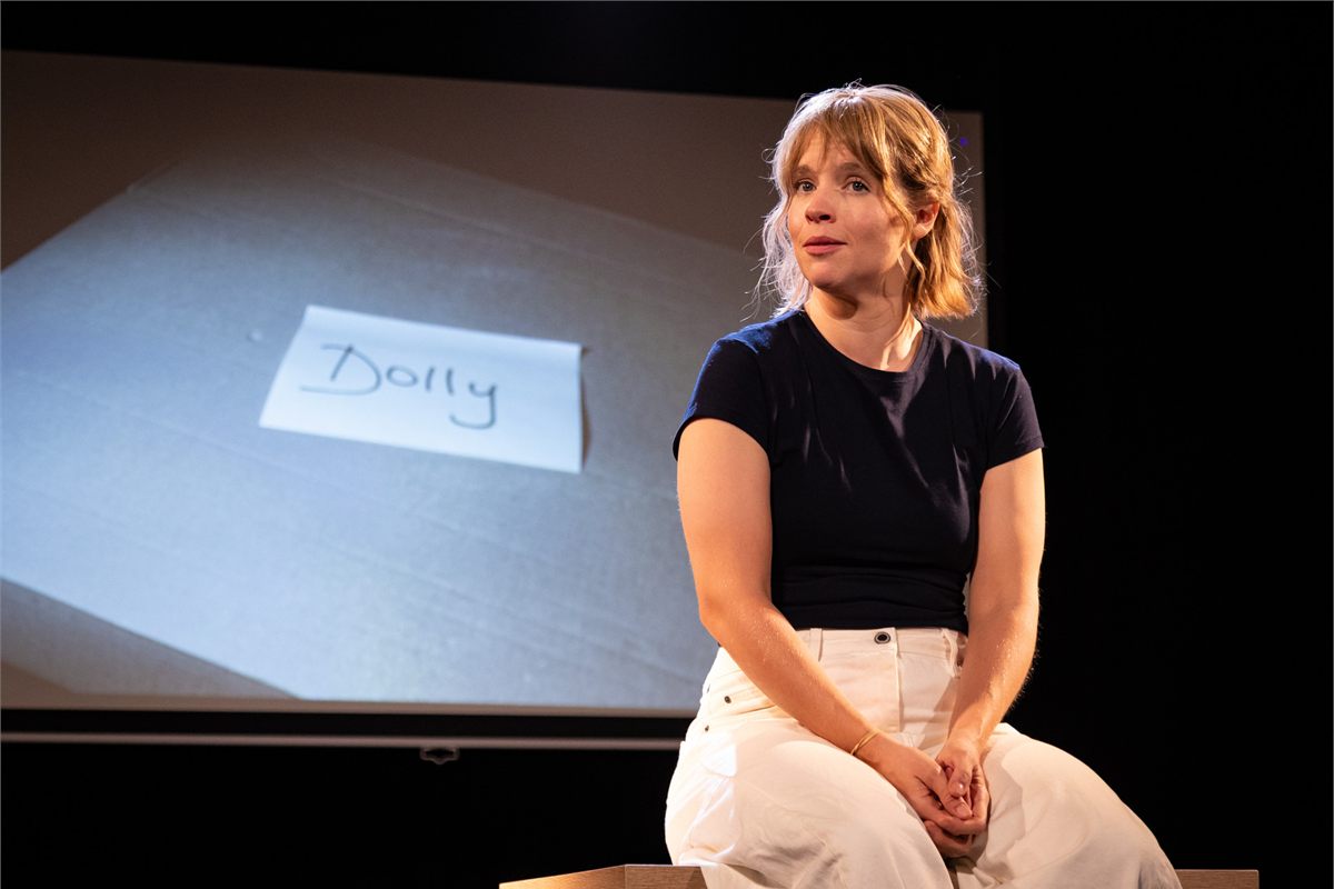 Jessica Clark seated in front of a projected card reading Dolly in Dolly at the Omnibus Theatre