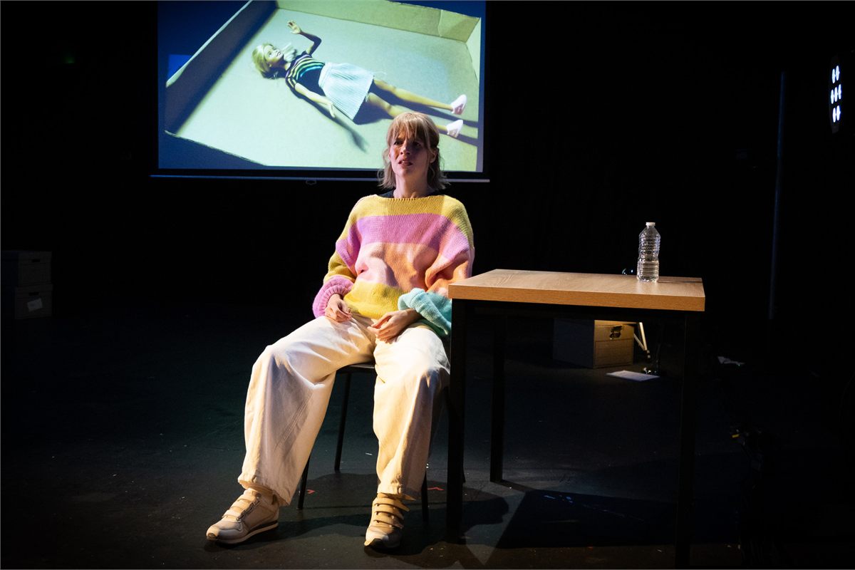 Jessica Clark seated beside a table beneath a projected image of a doll in Dolly at the Omnibus Theatre