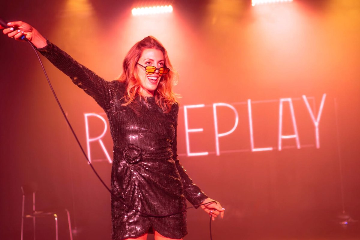 Hannah Reilly in a sequinned dress and sunglasses points a microphone towards the audience under red light in Roleplay Hannah Reilly in a sequinned dress and sunglasses points a microphone towards the audience under red light in Roleplay
