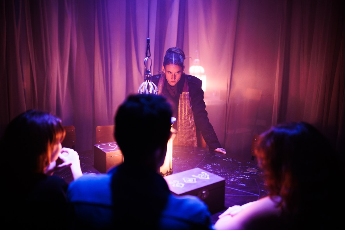 A performer looms over audience members seated at a candlelit table among purple curtains in Room 13 at Battersea Arts Centre A performer looms over audience members seated at a candlelit table among purple curtains in Room 13 at Battersea Arts Centre