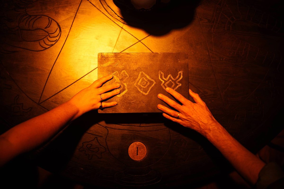 Two hands rest on a carved wooden box on a séance table lit by a lamp in Room 13 Two hands rest on a carved wooden box on a séance table lit by a lamp in Room 13