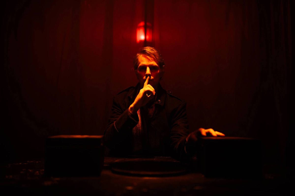 A performer raises a finger to his lips at a table under red light in Room 13 A performer raises a finger to his lips at a table under red light in Room 13