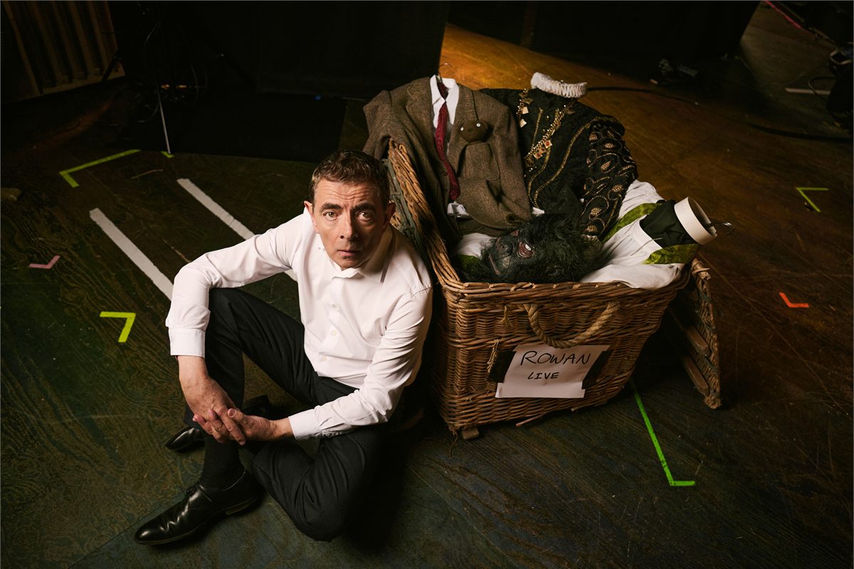 Rowan Atkinson seated on a stage floor beside a wicker costume hamper labelled Rowan Live