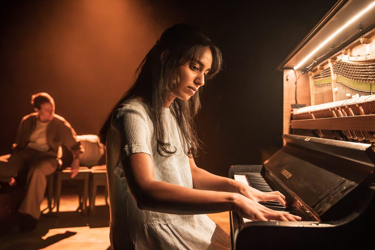 Kiah Arya as the young Noor at the piano in a small and quiet light. Photo by Marc Brenner
