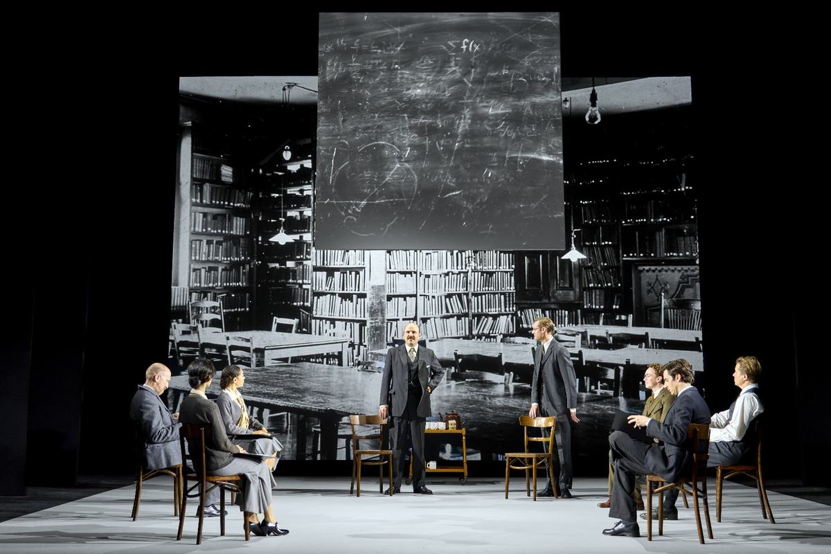 A lecture-room scene in The Standard of Living, with a blackboard flown in above the company. Photo by Manuel Harlan