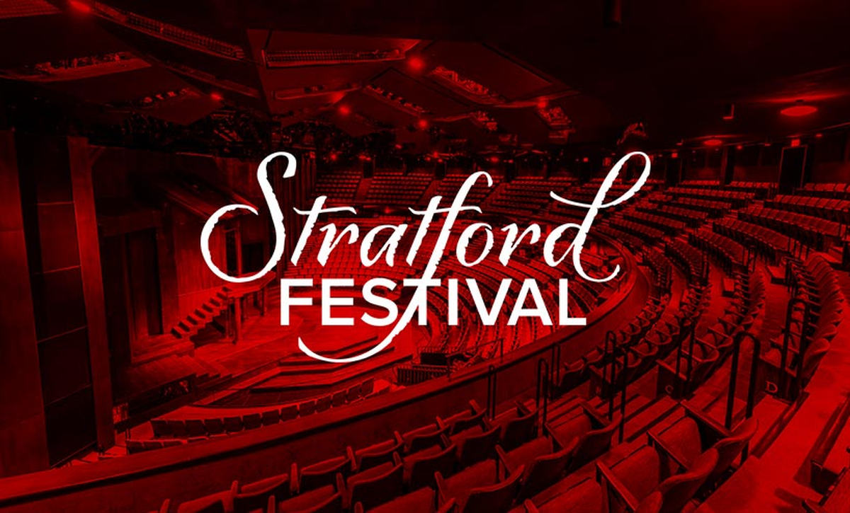 The auditorium of the Stratford Festival Theatre, its thrust stage and curved rows of empty seats lit in deep red, with the Stratford Festival logo across the centre