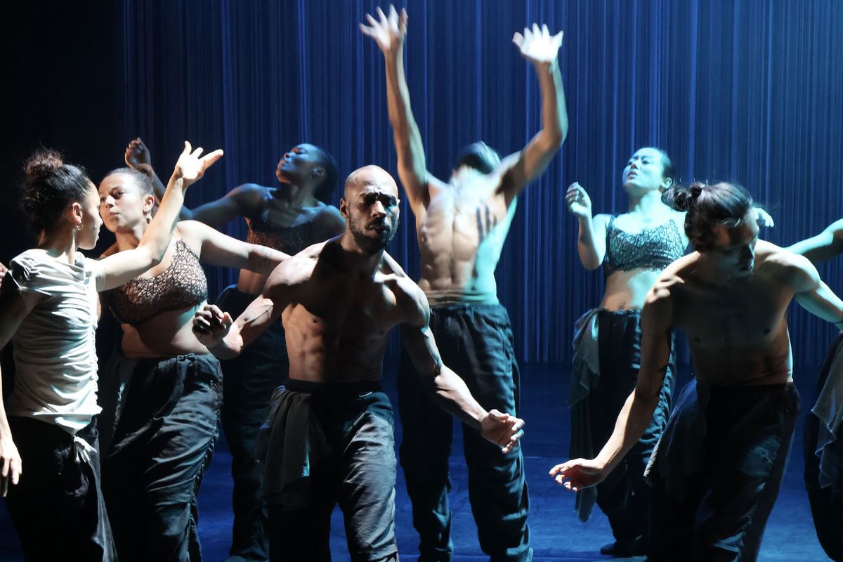 The company of Dickson Mbi Company's Tellus raising their arms under blue light at Sadler's Wells East