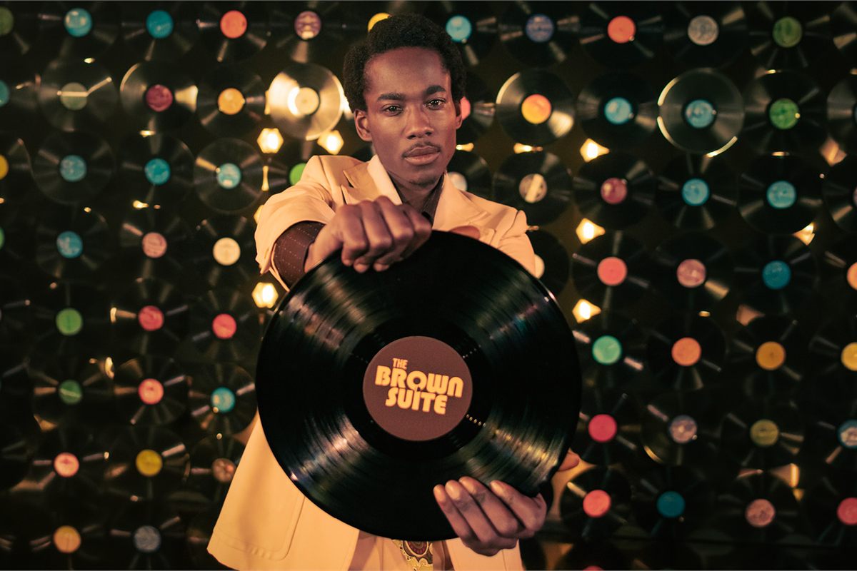 A performer in a cream suit holds out a vinyl record labelled The Brown Suite in front of a wall of records