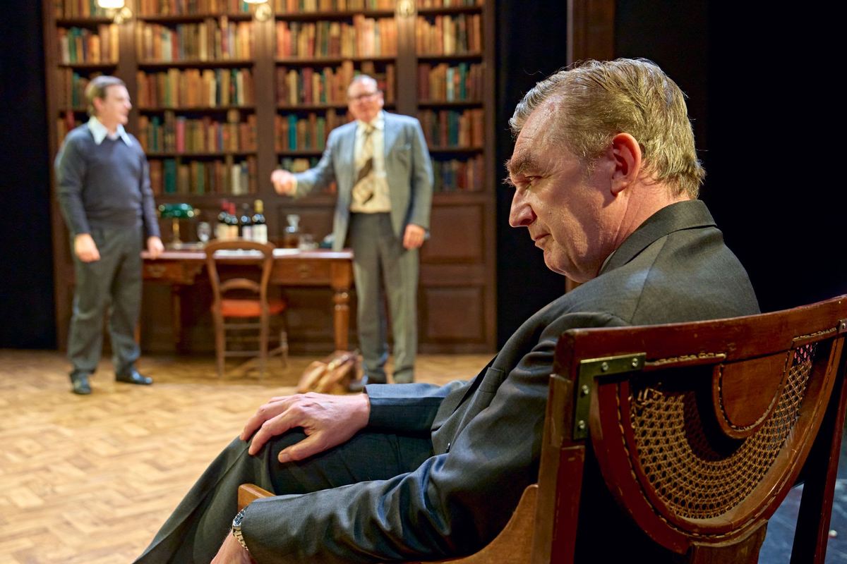 A seated figure in the foreground as the other two talk by the desk in The Gang of Three