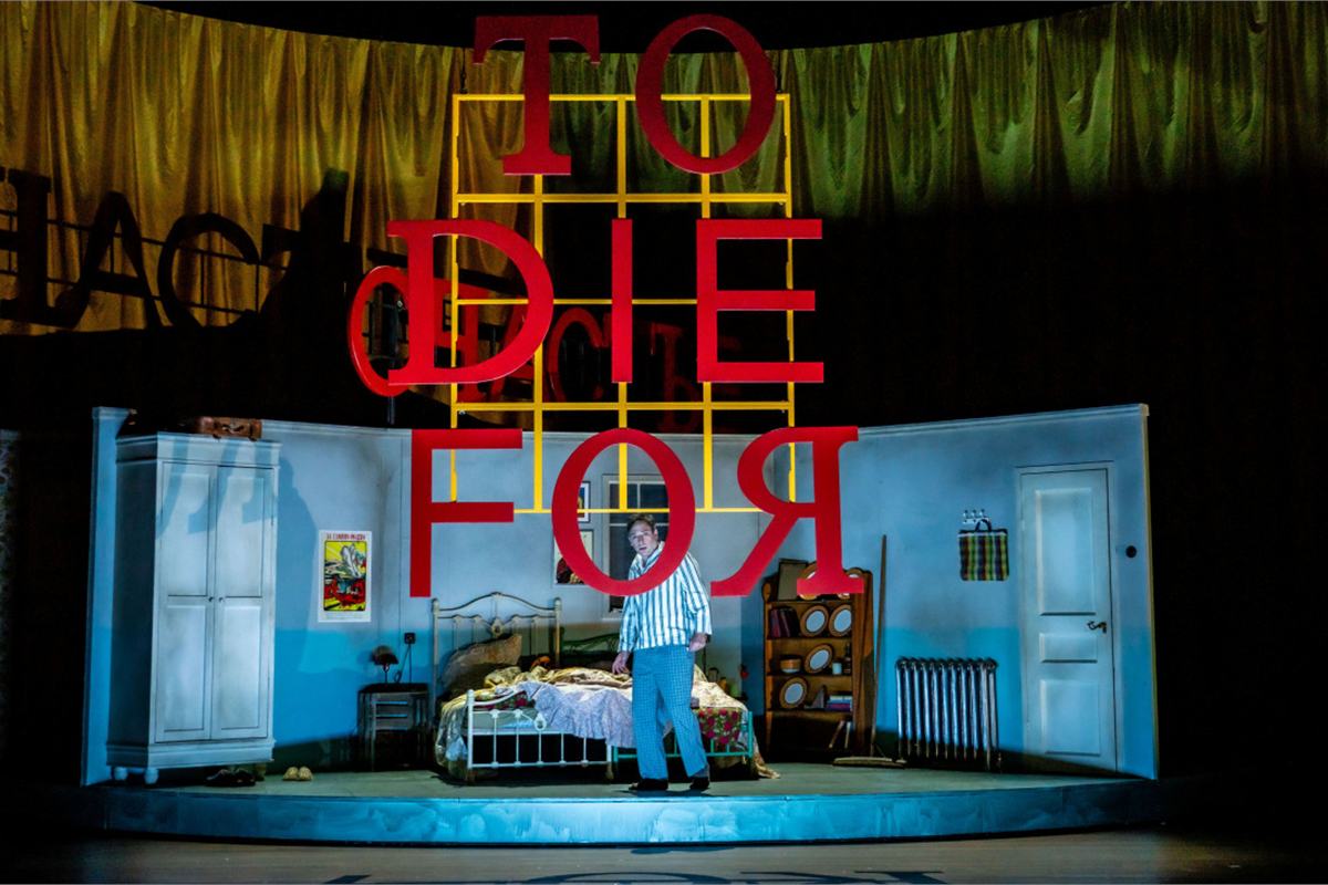 The set of To Die For [A Comedy] beneath its title in red neon, Nederlandse Reisopera's 2026 Dutch touring production