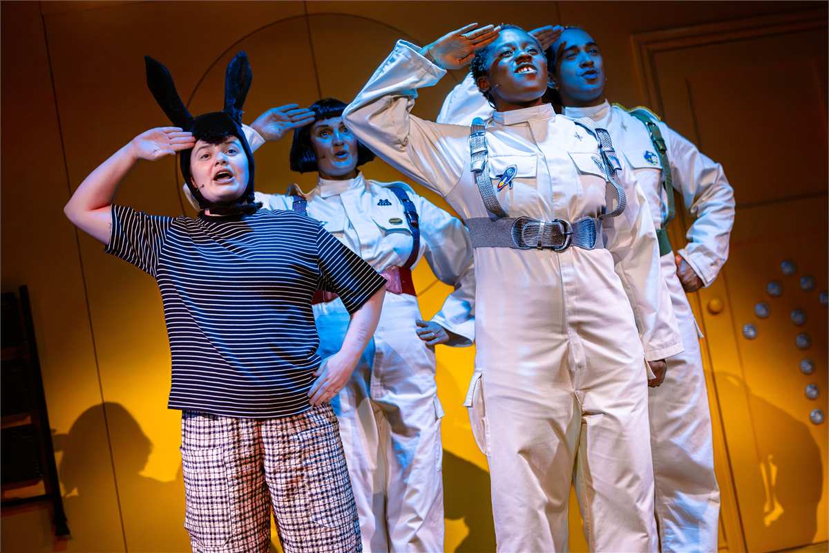 A donkey in a striped T-shirt salutes alongside three astronauts in white flight suits in Toto at Polka Theatre