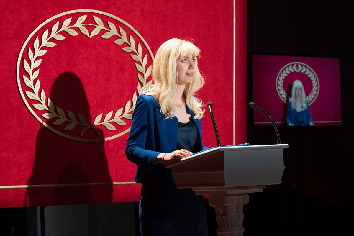 Kate O’Flynn as Iris at the lectern, with her broadcast on screen behind, in Triumph at the Almeida Theatre. Photo by Marc Brenner